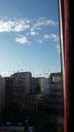 Buildings against sky seen through wet glass window