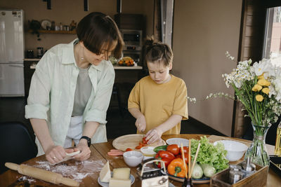 Smiling mother looking at son cutting tomato on table