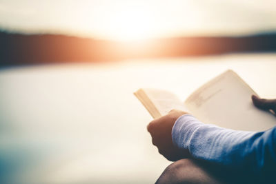 Midsection of person holding book against sky during sunset