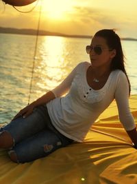 Young woman wearing sunglasses sitting on sea against sky during sunset