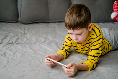 A teenage boy lies on the couch with a phone in his hands. the child plays at home in the phone. 