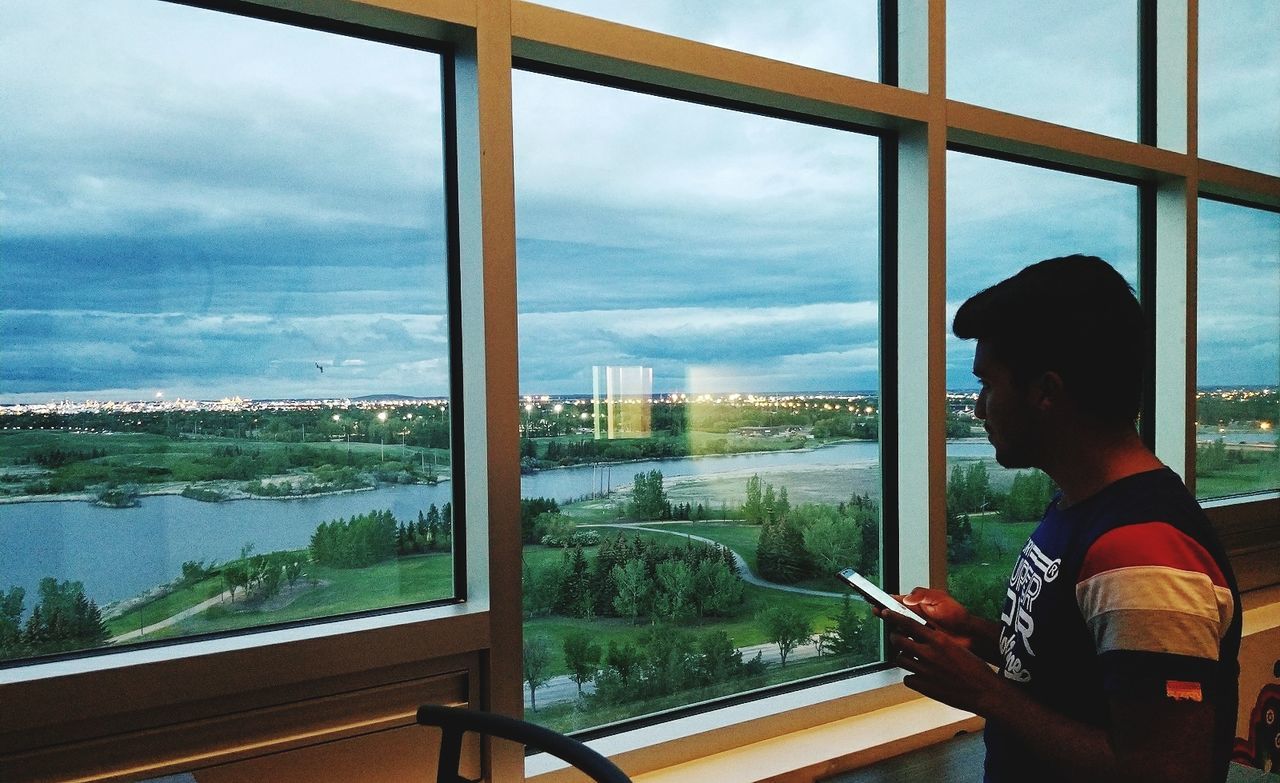 window, sky, glass - material, looking through window, cloud - sky, one person, real people, day, indoors, built structure, side view, looking at view, mountain, nature, tree, landscape, cityscape, lifestyles, architecture, standing, men, water, scenics, beauty in nature