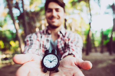Portrait of smiling man showing alarm clock while standing in forest