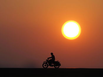 Silhouette man riding bicycle against orange sky