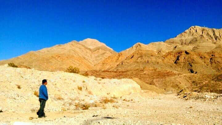 clear sky, blue, full length, lifestyles, leisure activity, mountain, men, rear view, landscape, rock formation, tranquility, rock - object, copy space, tranquil scene, nature, arid climate, desert, sunlight