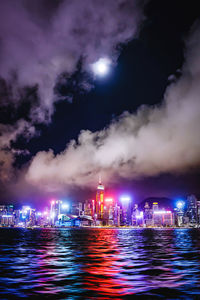 Illuminated buildings by sea against sky at night