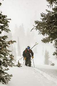 Man with skies walking on snow covered field in forest during snowfall