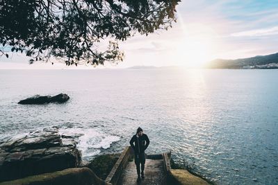 Rear view of man sitting on rock by sea against sky during sunset