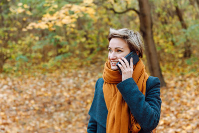 Short-haired blonde smiles as she talks on the phone in the fall outdoors