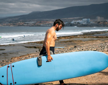 Side view of shirtless man holding surfboard at beach