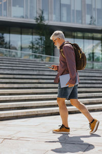 A senior businessman in a shirt and shorts climbs the steps to the office with a laptop