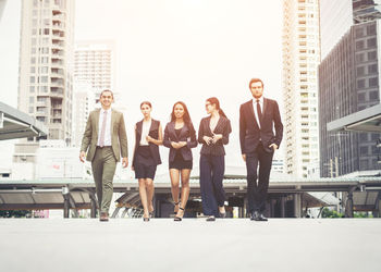 Portrait of young colleagues walking on footpath against buildings in city