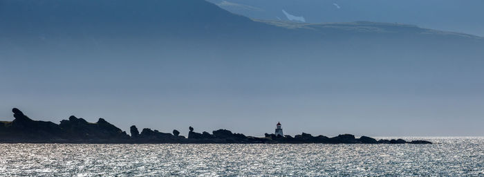 Distant view of lighthouse on rocks by sea against blue sky