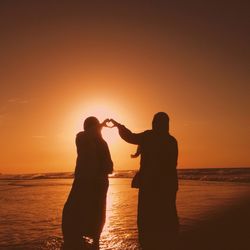 Silhouette women forming heart shape while standing in sea against sky during sunset