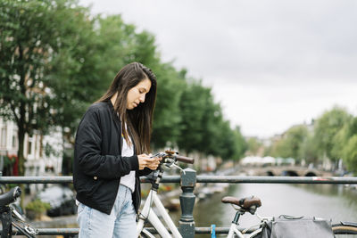 Side view of concentrated ethnic female traveler with long dark hair in casual clothes messaging on mobile phone while standing near parked bicycle on bridge during trip in amsterdam