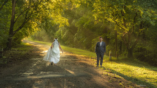 Portrait of bridegroom while bride walking on footpath amidst trees