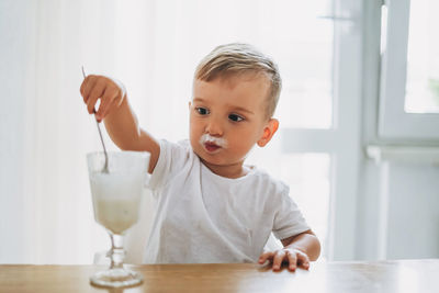 Charming concentrated little baby boy eating yogurt from glass at the bright kitchen at home