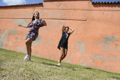 Two girls in summer dresses dancing in the park, against the backdrop of torange wall