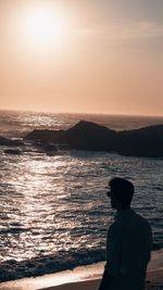 Rear view of man standing at beach against sky during sunset