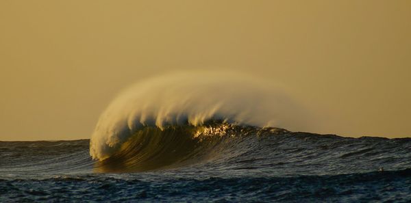 Sea waves splashing on shore against sky during sunset