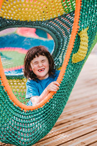 Adorable content little girl in t shirt smiling happily while relaxing with closed eyes inside of colorful knitted hammock in park