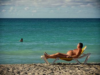 Side view of shirtless man relaxing on deck chair at beach against sky