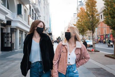 Laughing female best friends in protective masks walking along pavement in madrid and having fun during weekend