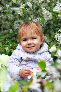 Happy smiling little baby girl in mother's hands on background of blooming garden with apple trees.