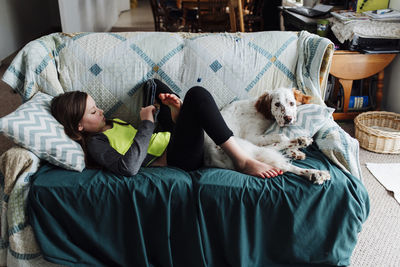 Girl using tablet computer while lying on sofa with dog at home