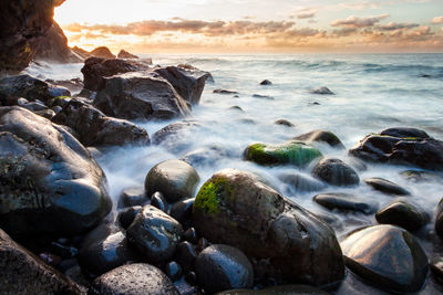 Waves splashing on rocks at beach against cloudy sky during sunset