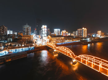 Illuminated bridge over river by buildings against sky at night