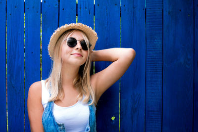Pretty teenage hipster girl smiling wearing straw hat and sunglasses outdoor over blue background.
