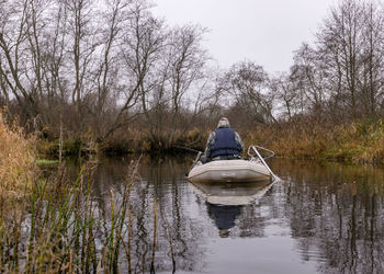 Fisherman in a white boat, river bank with bare trees and bushes, shore reflection in river water