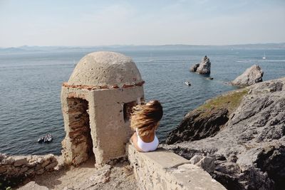 Rear view of woman sitting on retaining wall against sea at beach