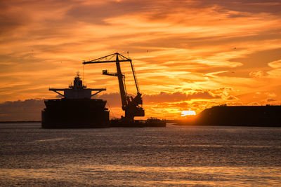 Silhouette ship and crane in river against orange sky