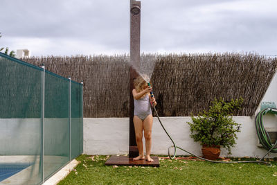 Cute little girl in swimsuit walking and watering green lawn from hose during summer holidays in countryside