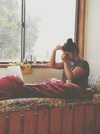 Woman trying jewelry while looking at laptop and sitting on bed by window at home