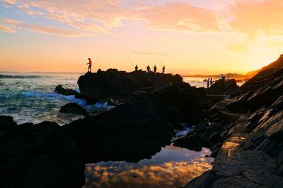 Friends fishing while standing on top of rock by sea during sunset
