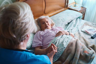 From above of nurse holding hand of aged female patient with gray hair lying on bed in bedroom at home