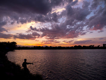 Silhouette person standing by lake against sky during sunset