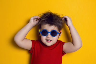 Cheerful baby boy in red t-shirt stands on yellow background in sunglasses