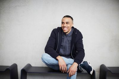 Smiling young student looking away while sitting on bench against wall in university