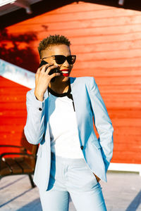 Cheerful african american female in trendy suit with hand in pocket talking on cellphone while looking away in sunny town
