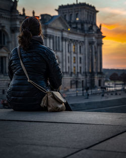 Rear view of woman sitting by historic building at sunset