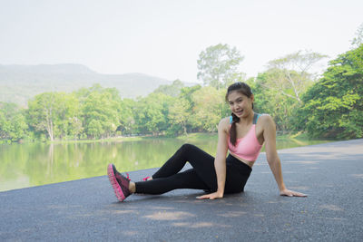 Full length portrait of young woman sitting on road by river at park