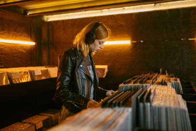 Side view of young female with headphones choosing vinyl record while standing near counter with collection of musical albums in store with glowing lamps