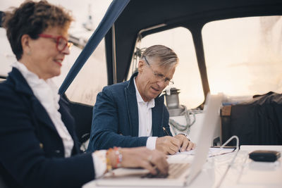 Senior man writing on paper while sitting by woman using laptop in yacht