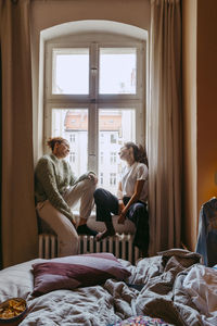 Young female friends talking while sitting at window in bedroom