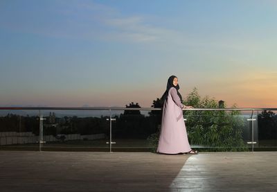 Woman standing by railing against sky during sunset