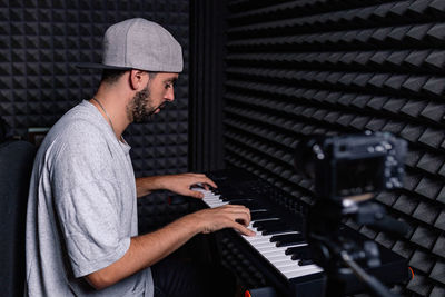 Side view of skilled male musician playing electric piano and recording video on camera in acoustic room with soundproof foam walls